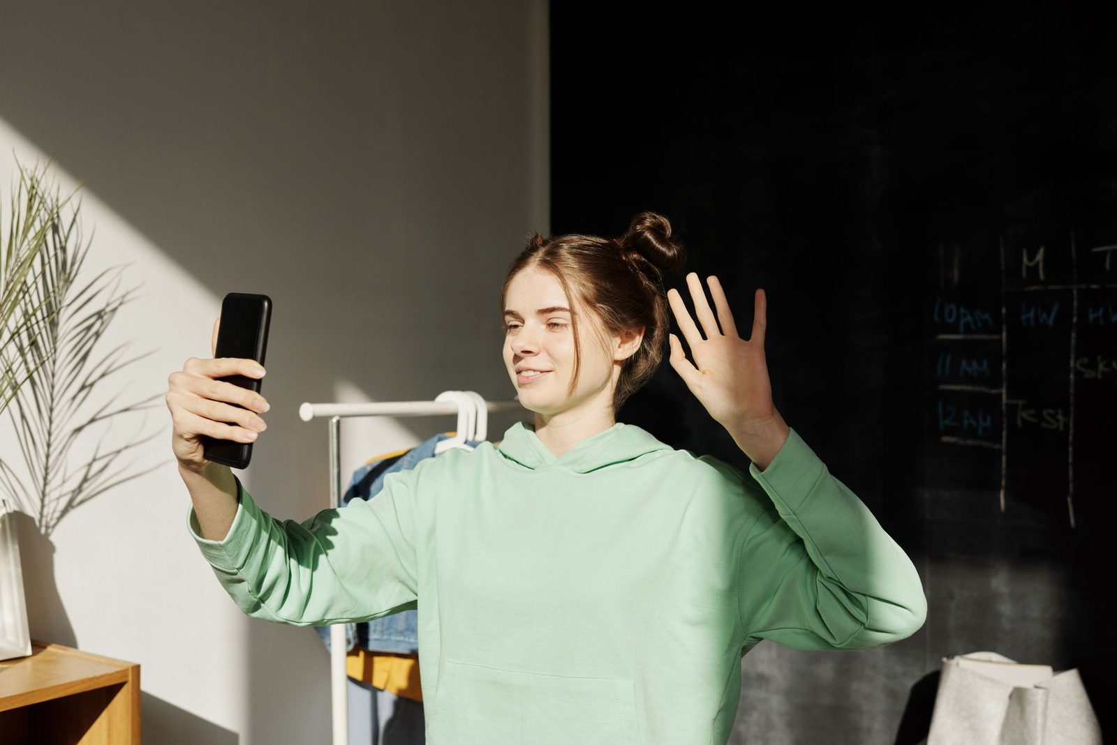 Yeşil sweatshirt giymiş genç kadın, akıllı telefonundan görüntülü görüşmede el sallıyor
