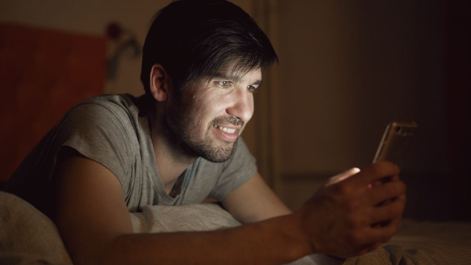 Homme souriant allongé dans son lit la nuit, regardant l'écran de son smartphone tenu à la main