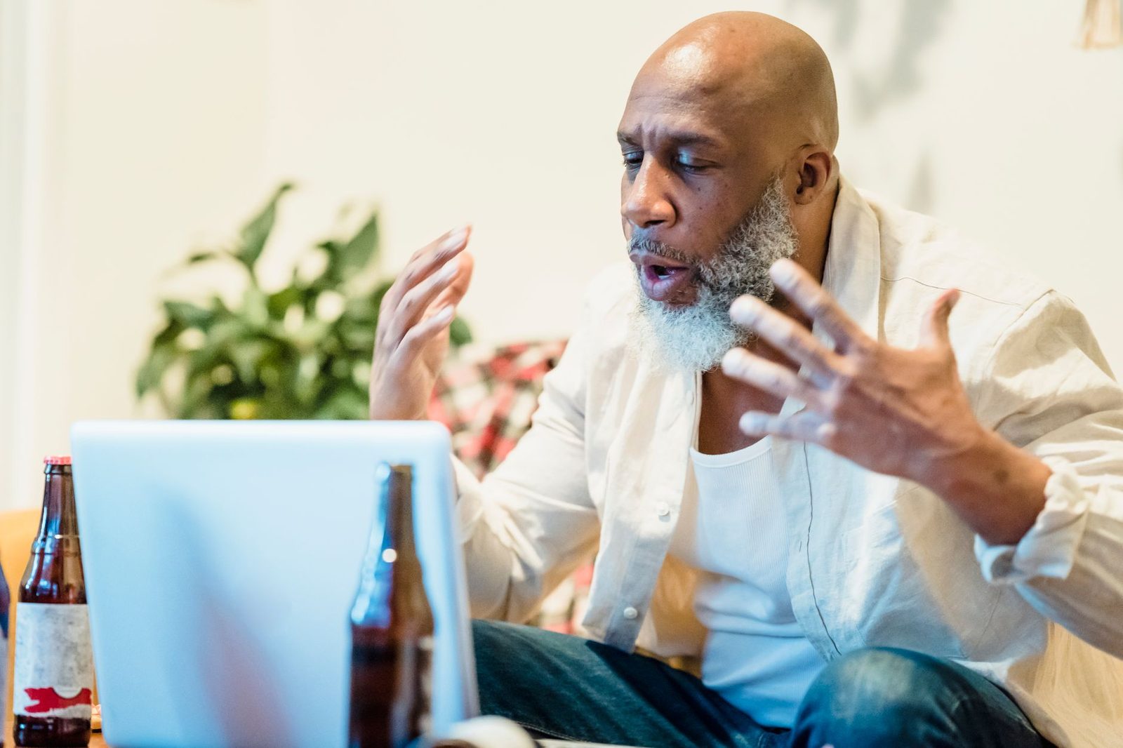 Homme barbu assis devant un ordinateur portable, gesticulant pendant une conversation vidéo, bouteilles sur la table
