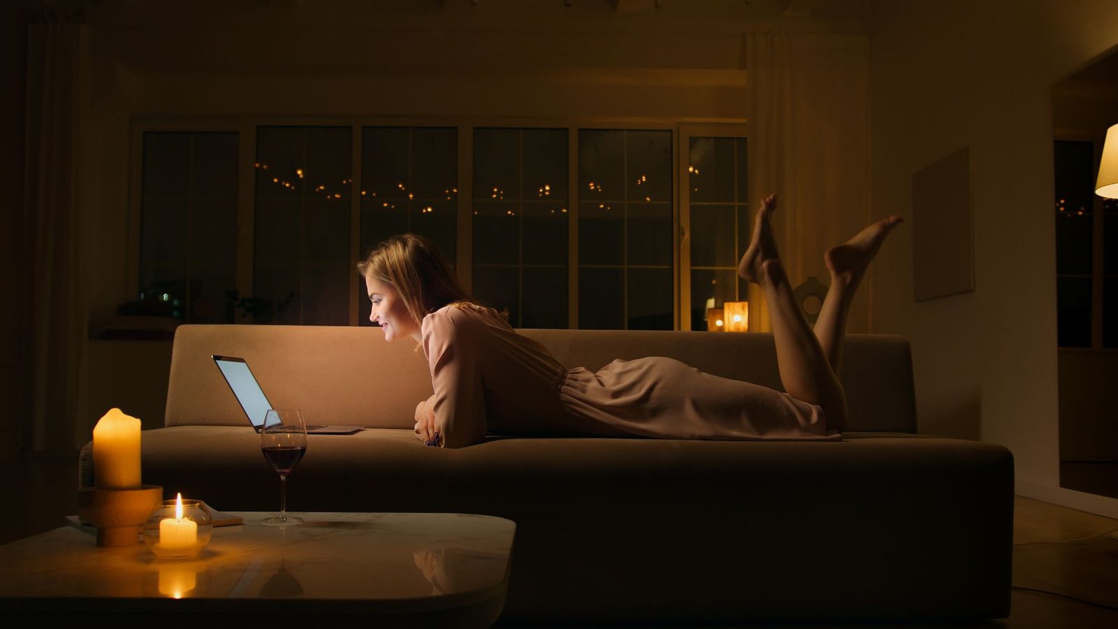Jonge vrouw ligt 's avonds op een bank, glimlachend voor haar laptop, met een glas wijn en kaarsen