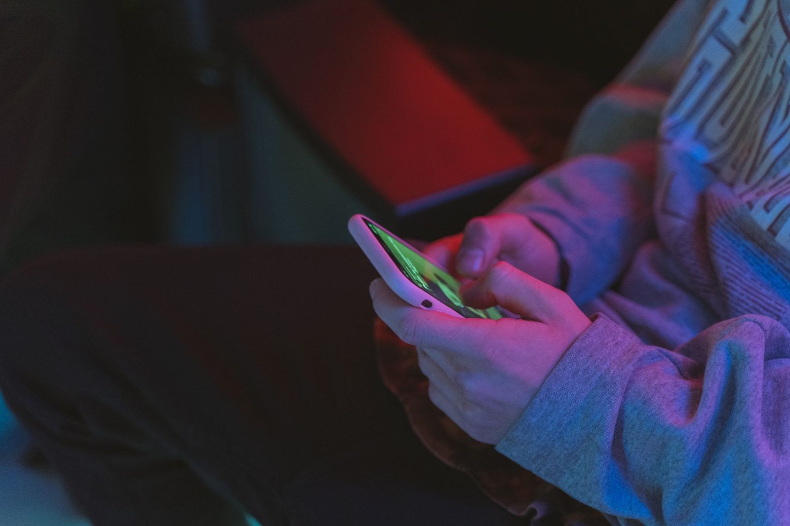 Handen van een persoon die een smartphone met verlicht scherm vasthoudt, in een donkere kamer met rode en blauwe lichten