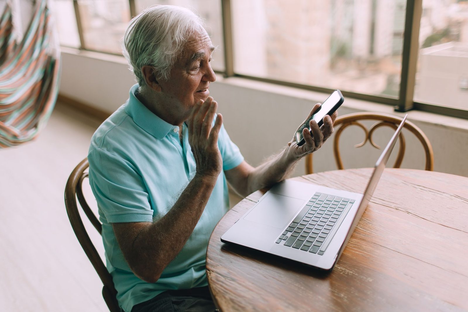 Oudere man in turquoise poloshirt zwaait naar een smartphone en een laptop