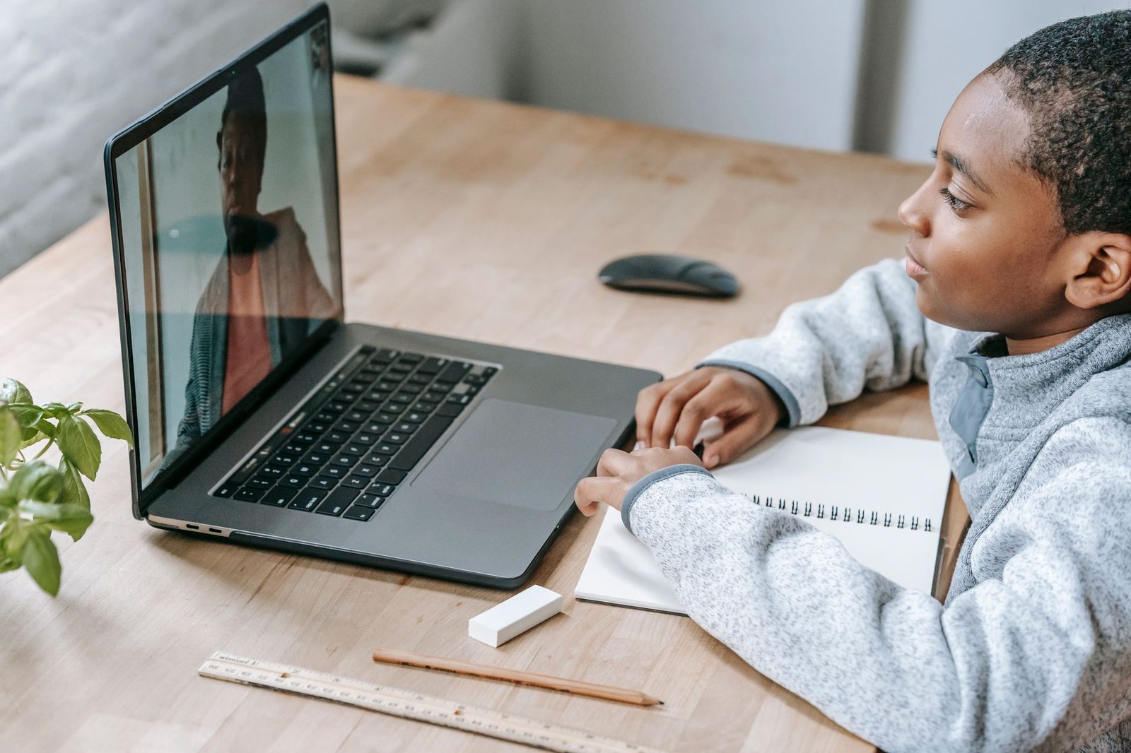 Jeune garçon en sweat gris devant un ordinateur portable pendant un appel vidéo, cahier et crayon sur la table