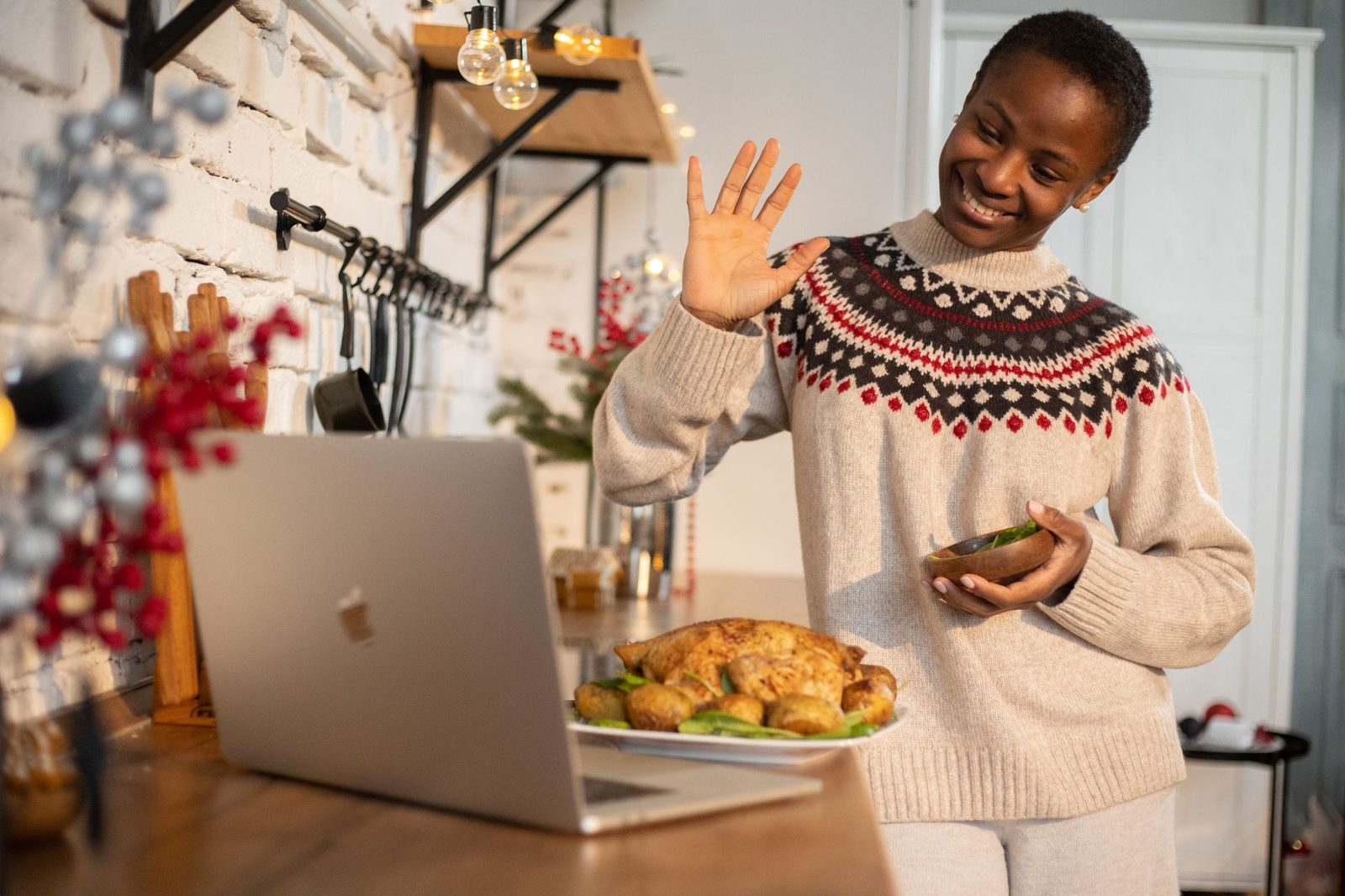 Mujer sonriente saludando con la mano frente a un ordenador portátil en una cocina decorada por Navidad