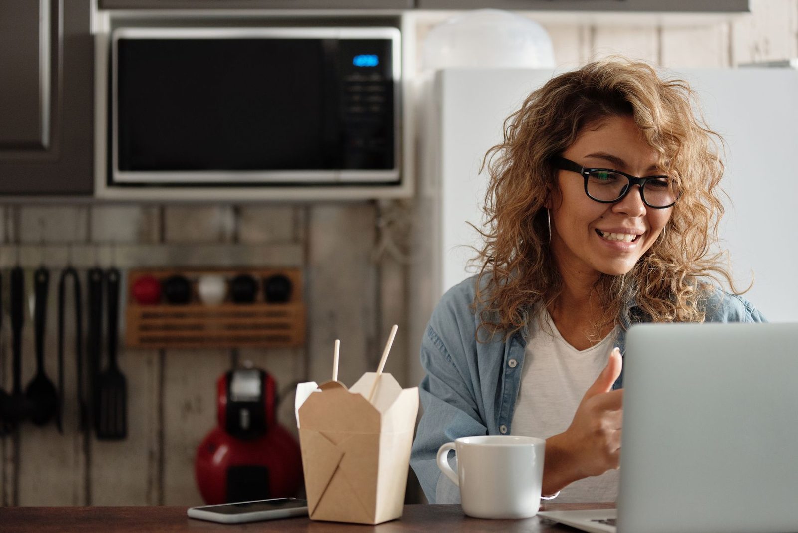 Giovane donna con occhiali sorridente davanti a un computer portatile in una cucina, tazza e piatto da asporto