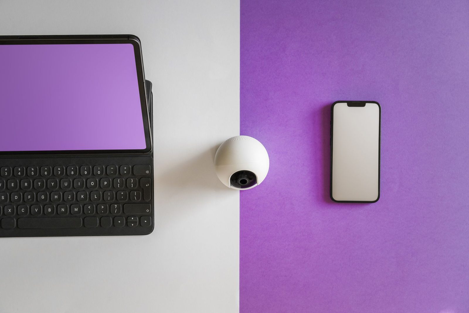 Tablet dengan keyboard, webcam putih, dan ponsel pintar diletakkan di atas latar putih dan ungu, dilihat dari atas