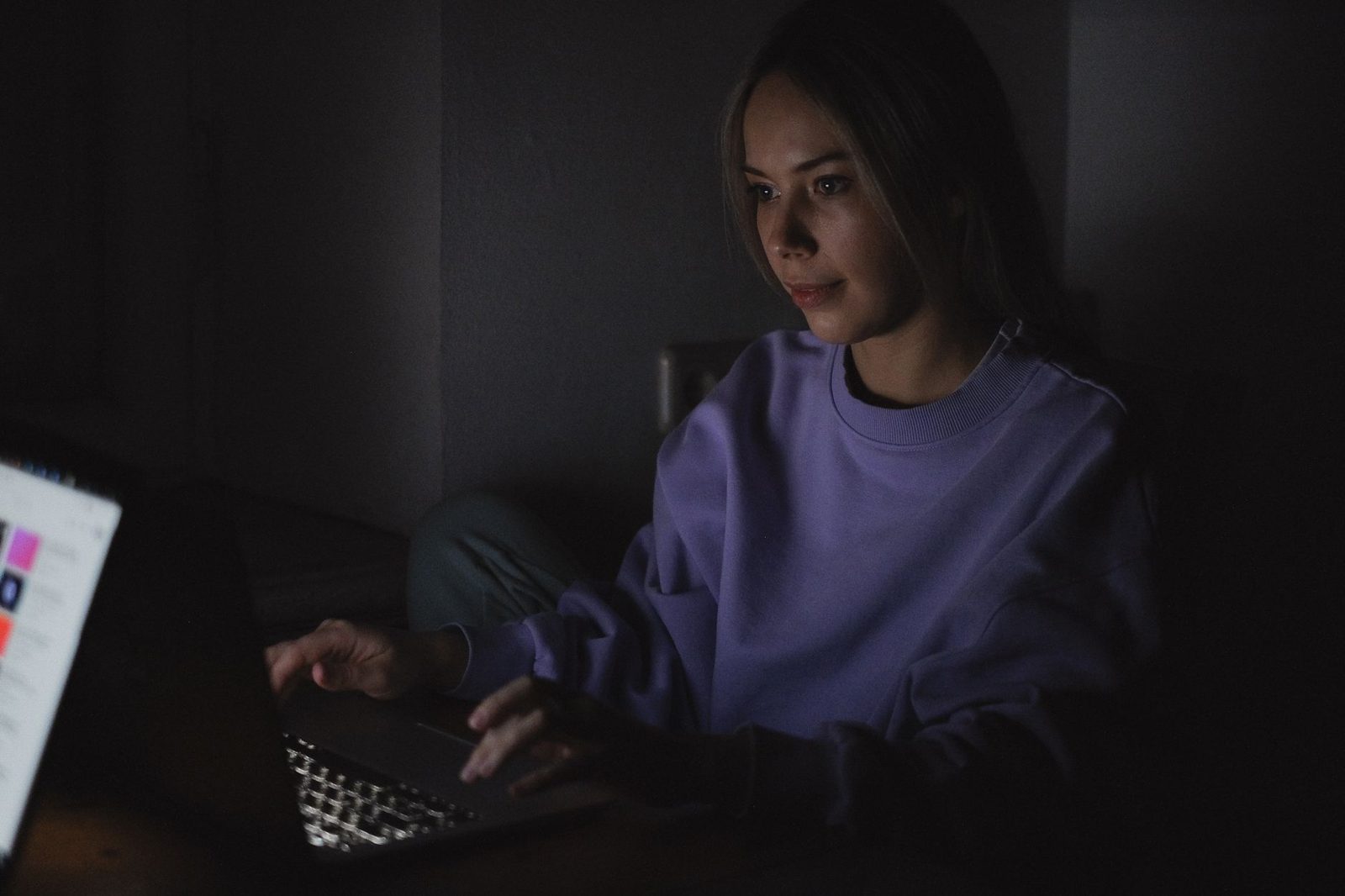 Young woman in a purple hoodie in front of a laptop, her face lit by the screen in a dark room