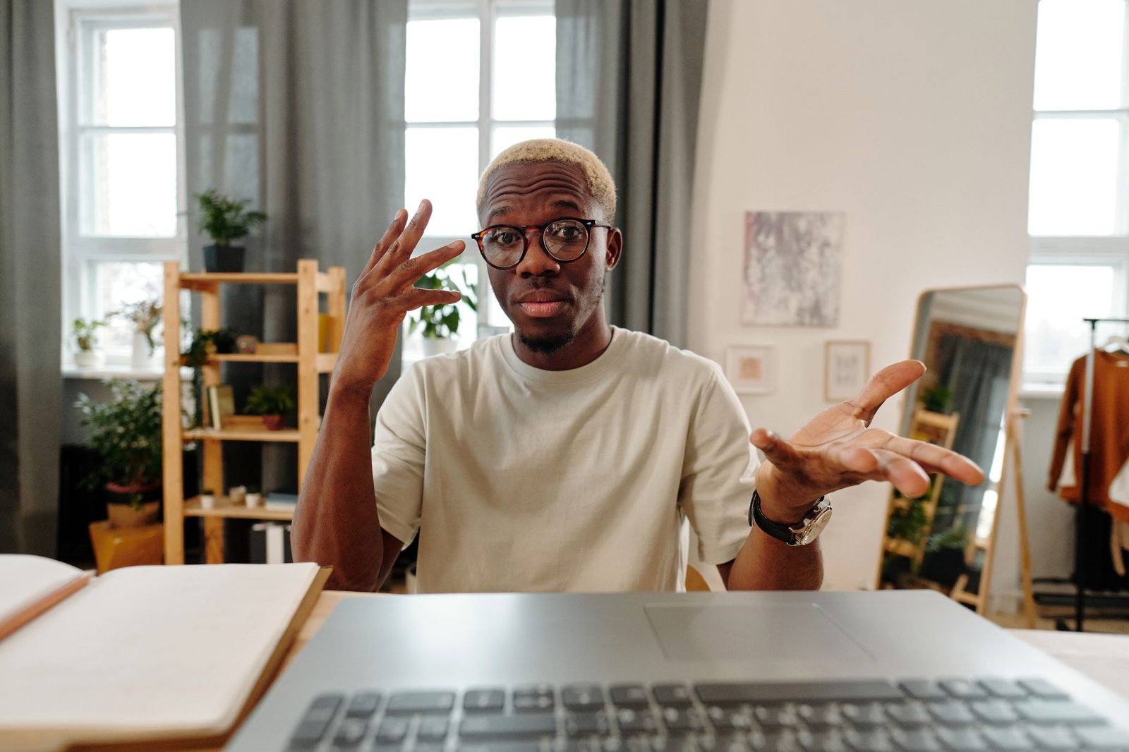 Man in glasses sitting in front of a laptop, hands raised, mid-video call at home