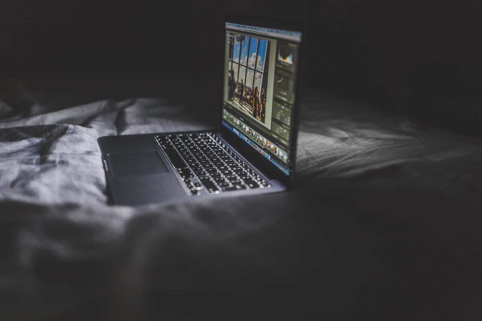 Open laptop switched on, sitting on a bed with rumpled sheets in a dark bedroom
