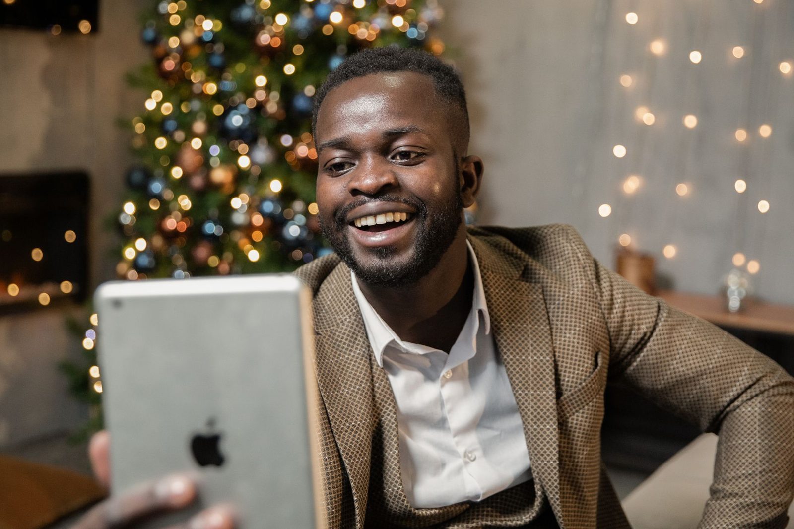 Homme souriant en costume lors d'un appel vidéo sur tablette, devant un sapin de Noël illuminé