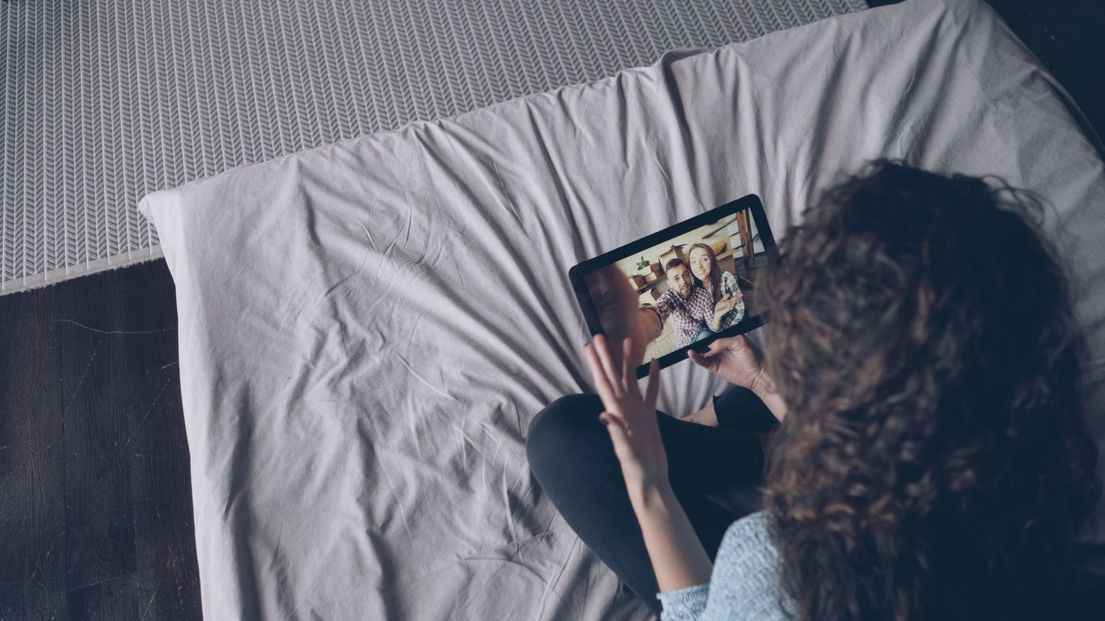 Junge Frau sitzt auf einem Bett und winkt während eines Videoanrufs auf einem Tablet