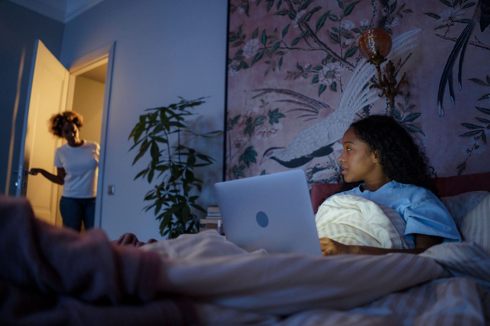 Adolescente sdraiata nel letto di notte davanti a un portatile, una donna sulla porta